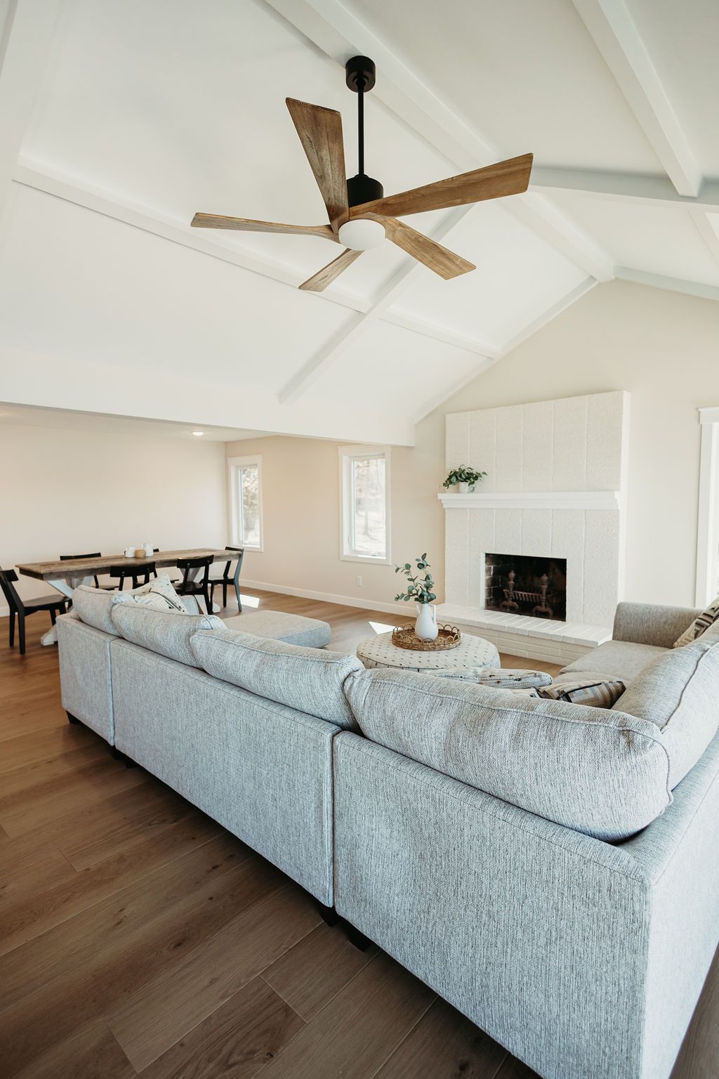 Bright living room with gray sectional, wood floors, ceiling fan, dining area, and fireplace