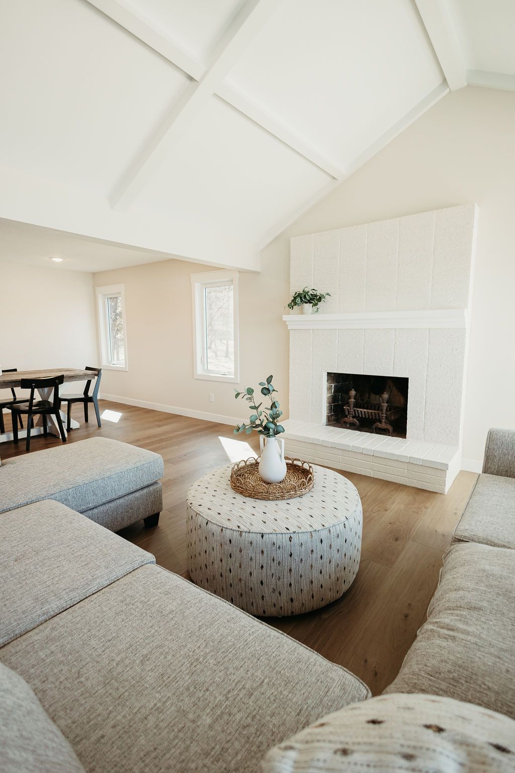 Bright living room with white vaulted ceiling, fireplace, gray sectional, round ottoman, and wood floors