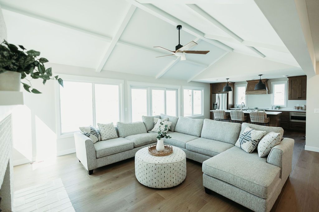 Bright open living room with white walls, light sofas, round ottoman, and large windows