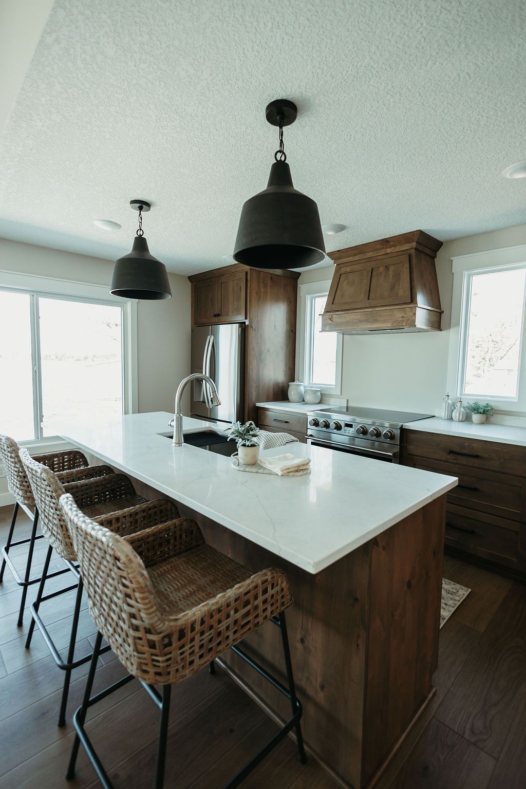 Modern kitchen with a white island, woven bar stools, wood cabinets, and hanging black pendant lights
