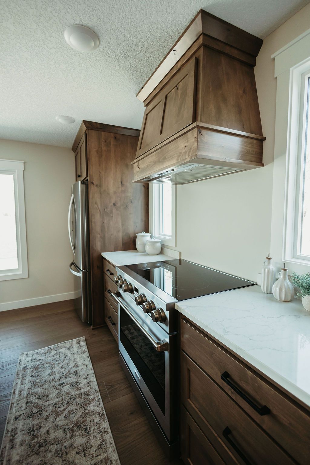 Narrow modern kitchen with wood cabinets, white countertops, stainless fridge, and range by windows