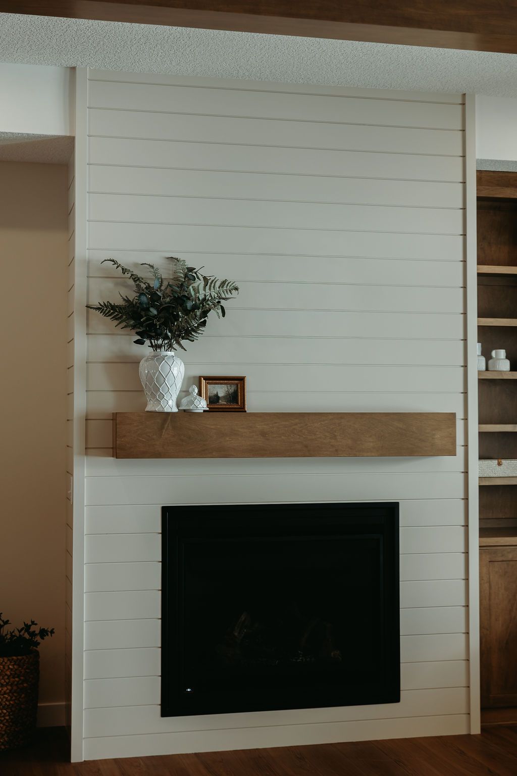 Fireplace with white shiplap, wooden mantel, and black firebox, decorated with greenery and small art.