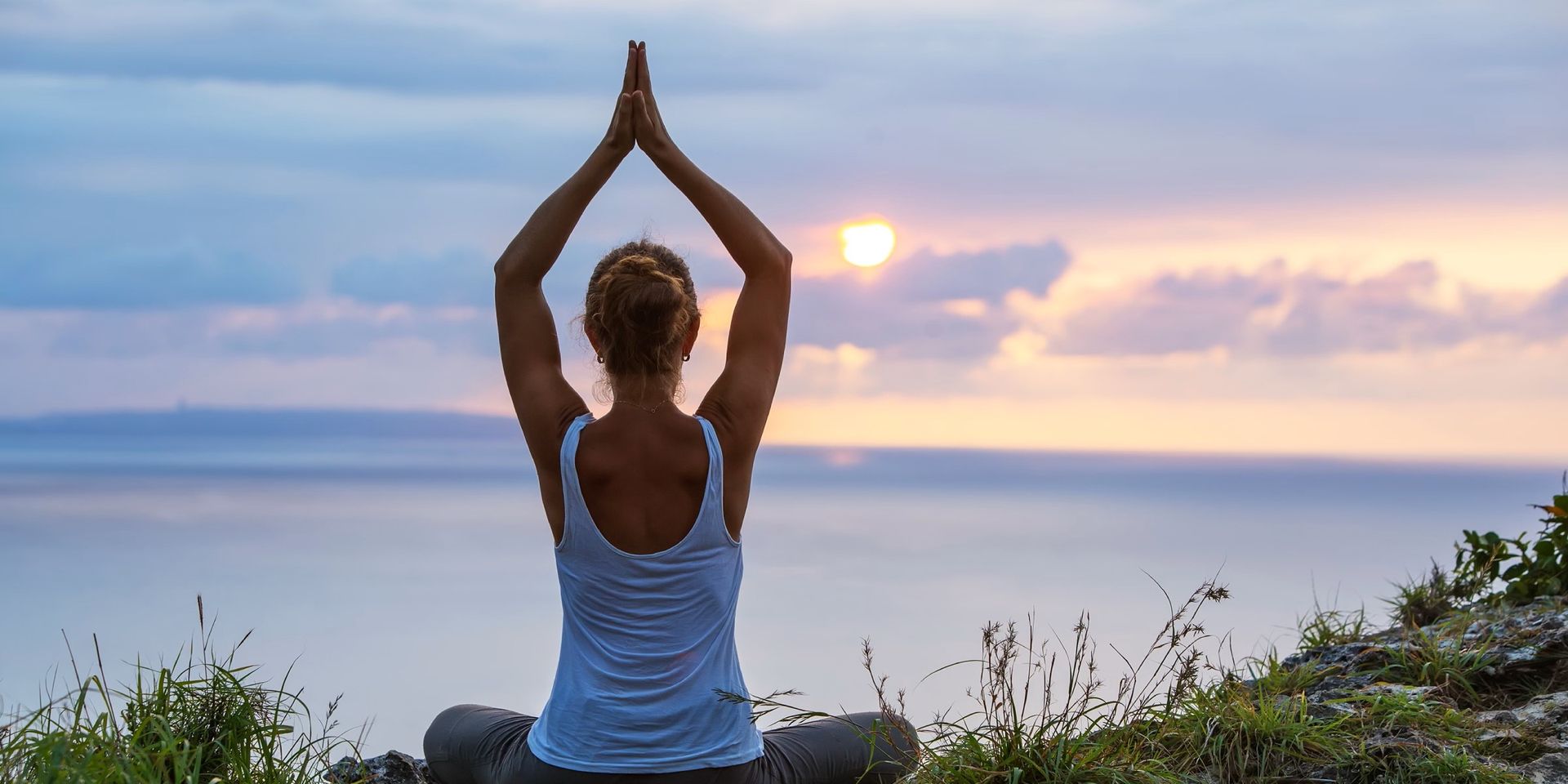 A woman is sitting in a yoga pose on top of a hill overlooking the ocean at sunset.