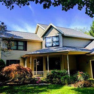 Two-story beige house with a porch, windows, and green lawn under a blue sky with trees.