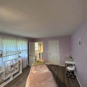 Massage room with lilac walls, a massage table, and a window with curtains.