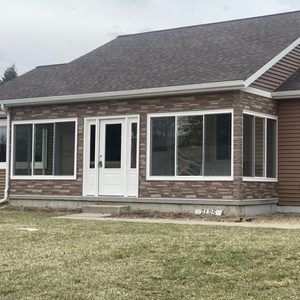 A house with stone veneer siding, large windows, a white front door, and a brown shingled roof on a grassy lawn.