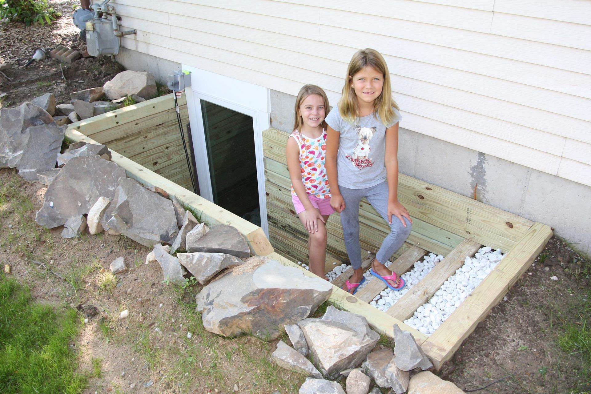 Two girls stand in a newly built window well. Wooden frame, white gravel, large rocks around it.