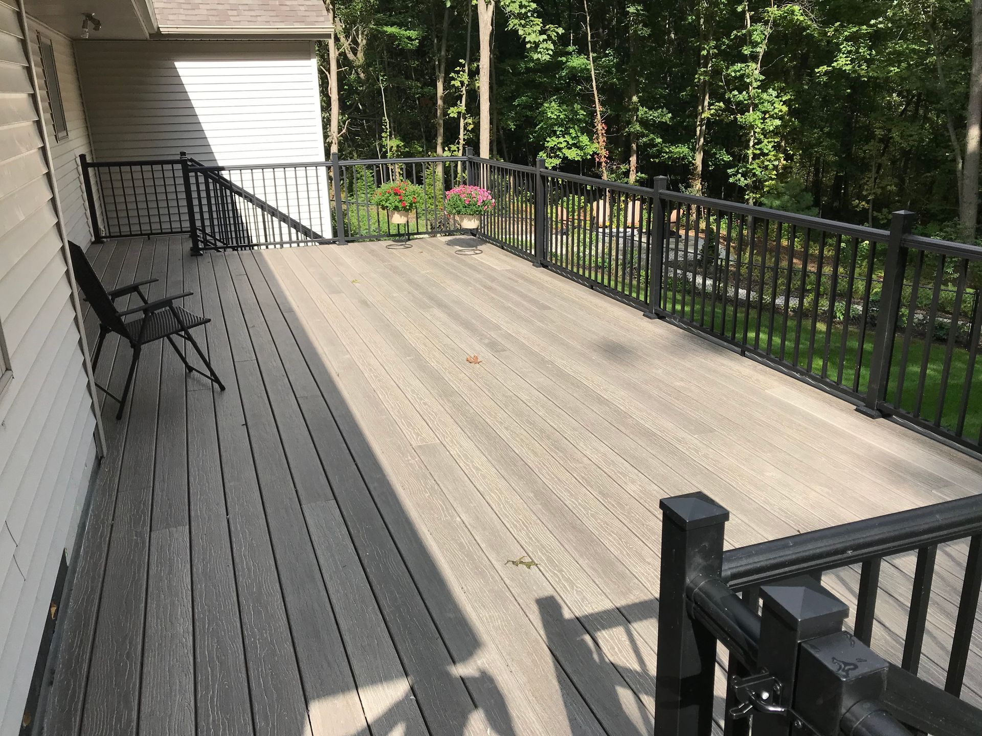 Wooden deck with black railings and chairs beside a house, overlooking trees and a yard