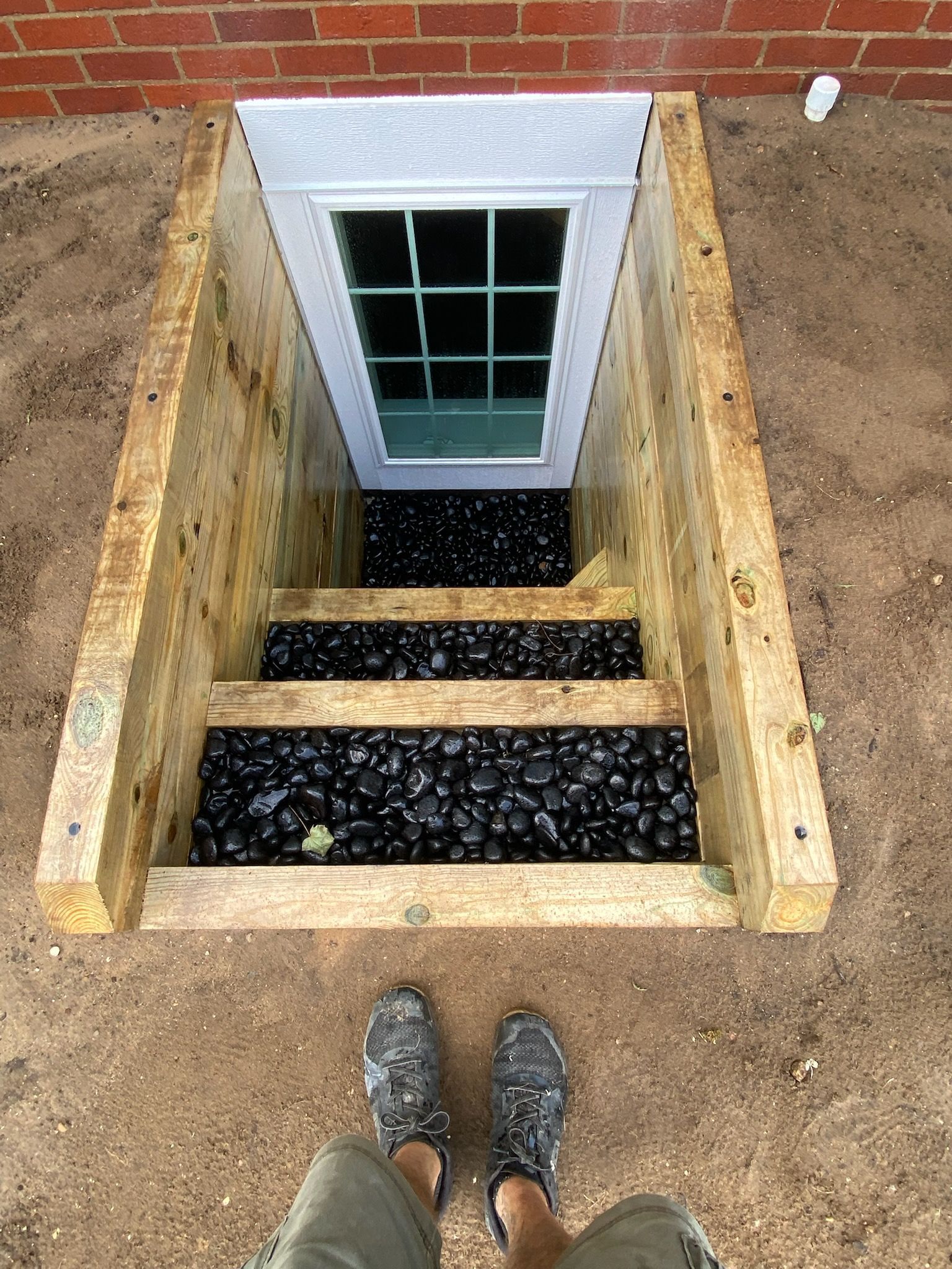 Basement window well with wooden steps and gravel, viewed from above over dirt ground.