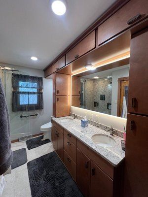 Bathroom with wood cabinets, granite countertop, large mirror with lighting, and a shower visible in the mirror.