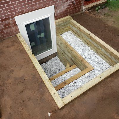 Wooden foundation framing around a basement window with gravel fill, beside a brick wall outdoors