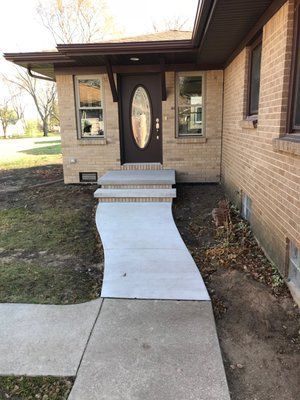 A concrete walkway curves toward a house's front door with steps. Brick facade, brown door.