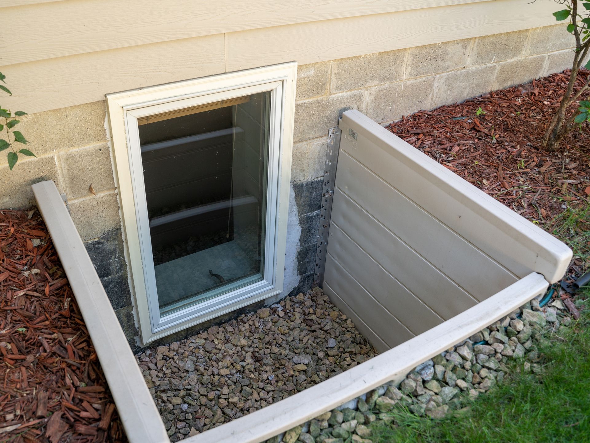 Basement window well with gravel, concrete walls, and a small exterior window beside a foundation wall