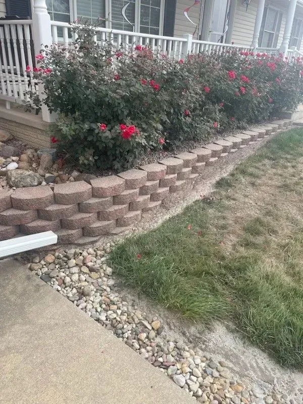 A retaining wall of tan concrete blocks borders a garden bed with red rose bushes in front of a white porch railing.