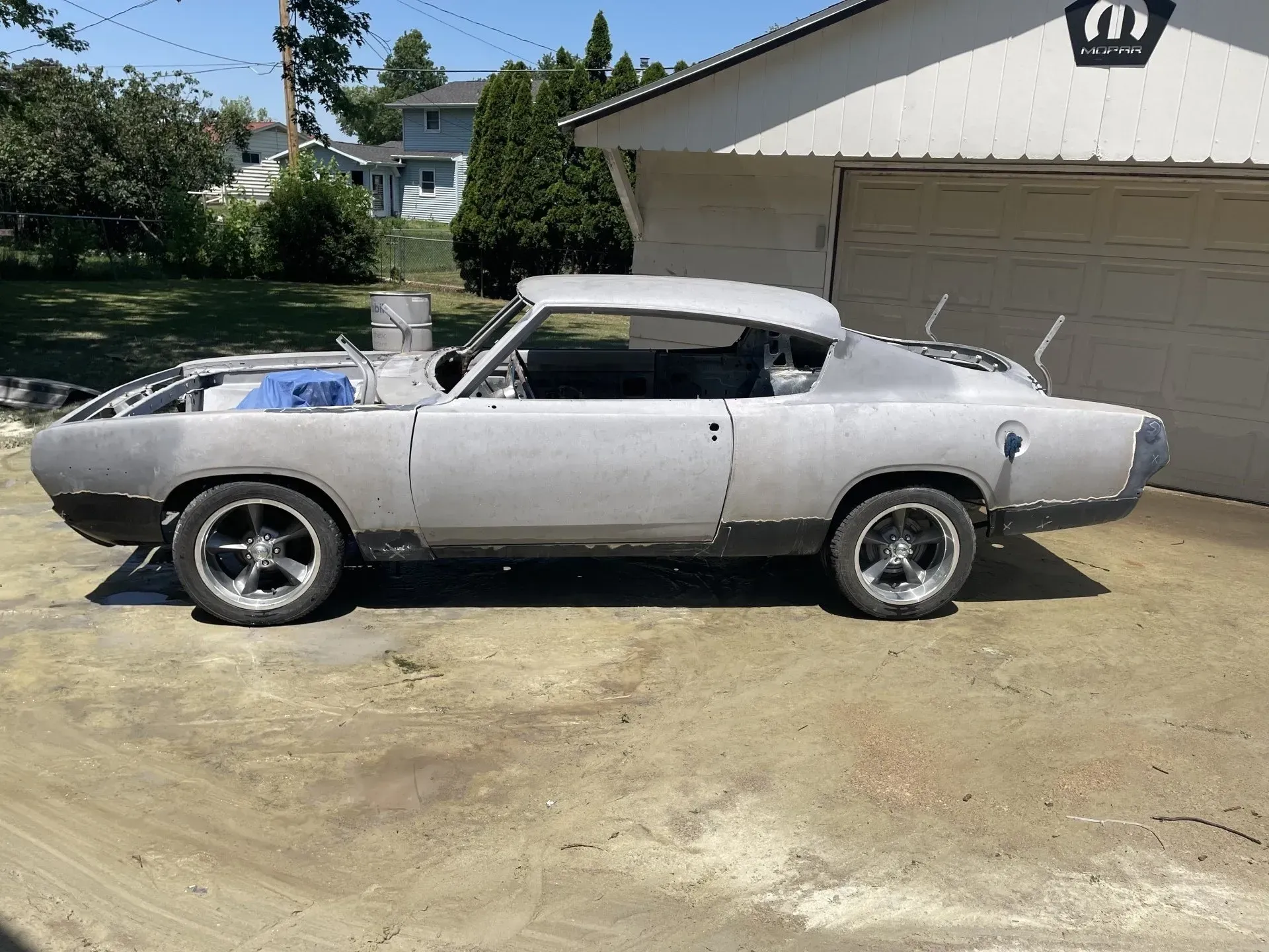 A grey, primer-coated classic car frame sits parked on a gravel driveway in front of a Mopar garage.