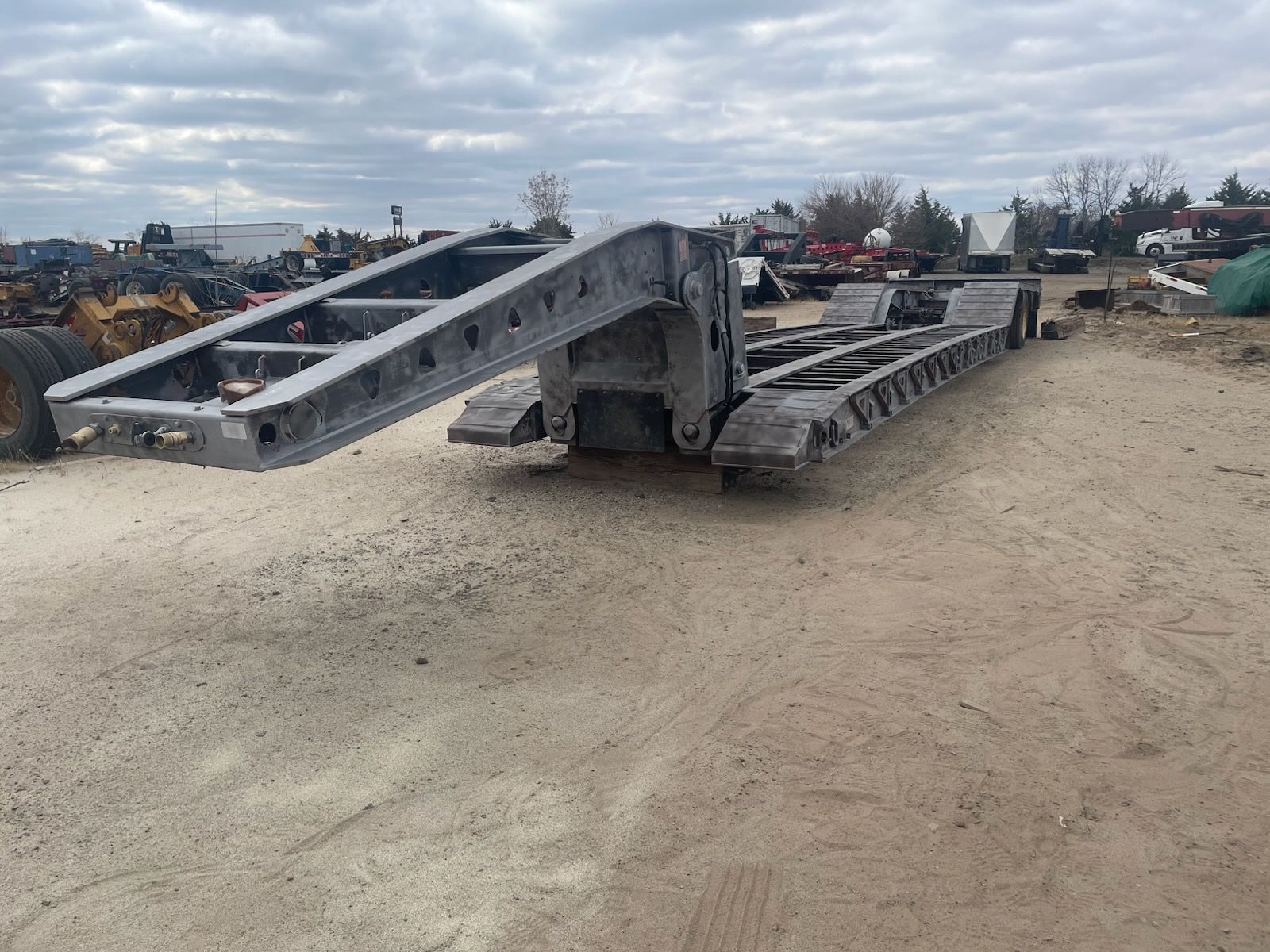 A gray, heavy-duty detachable gooseneck trailer sits on a gravel lot under a cloudy sky.