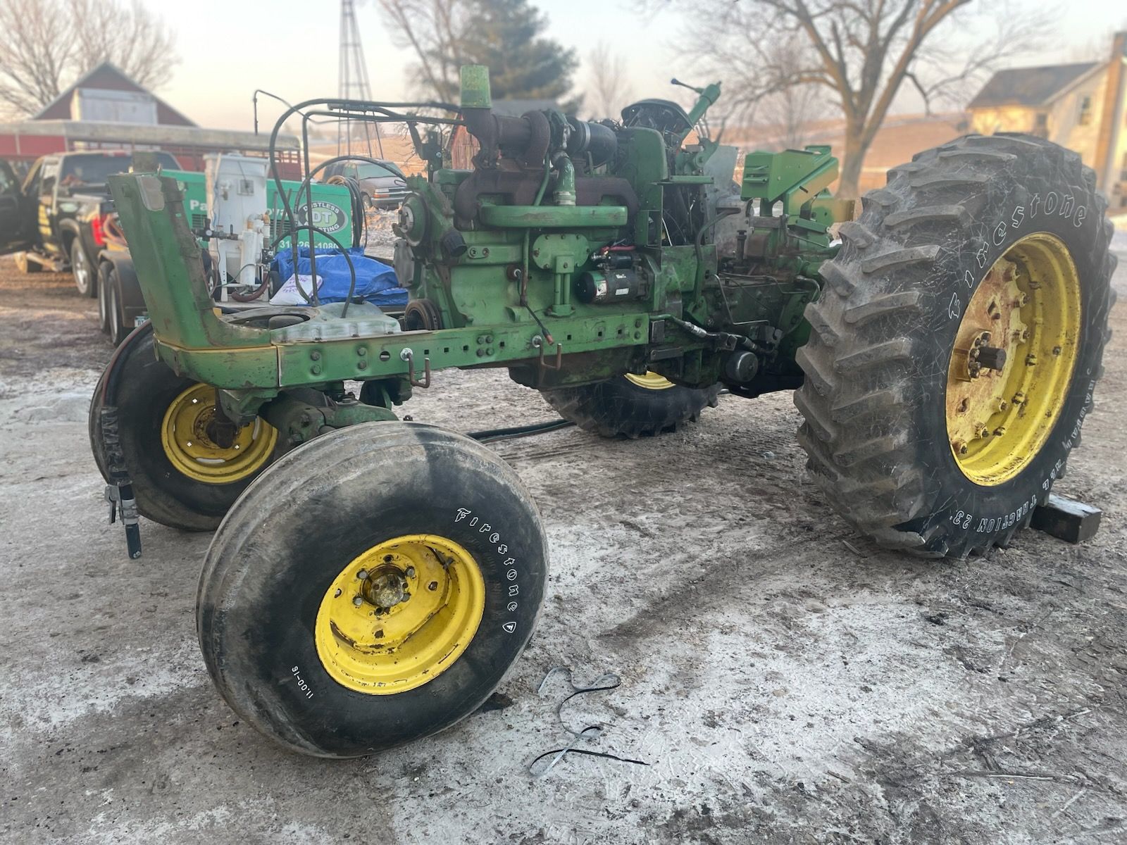 A partially disassembled green tractor parked on a dirt lot, featuring a large rear tire and a smaller front tire.