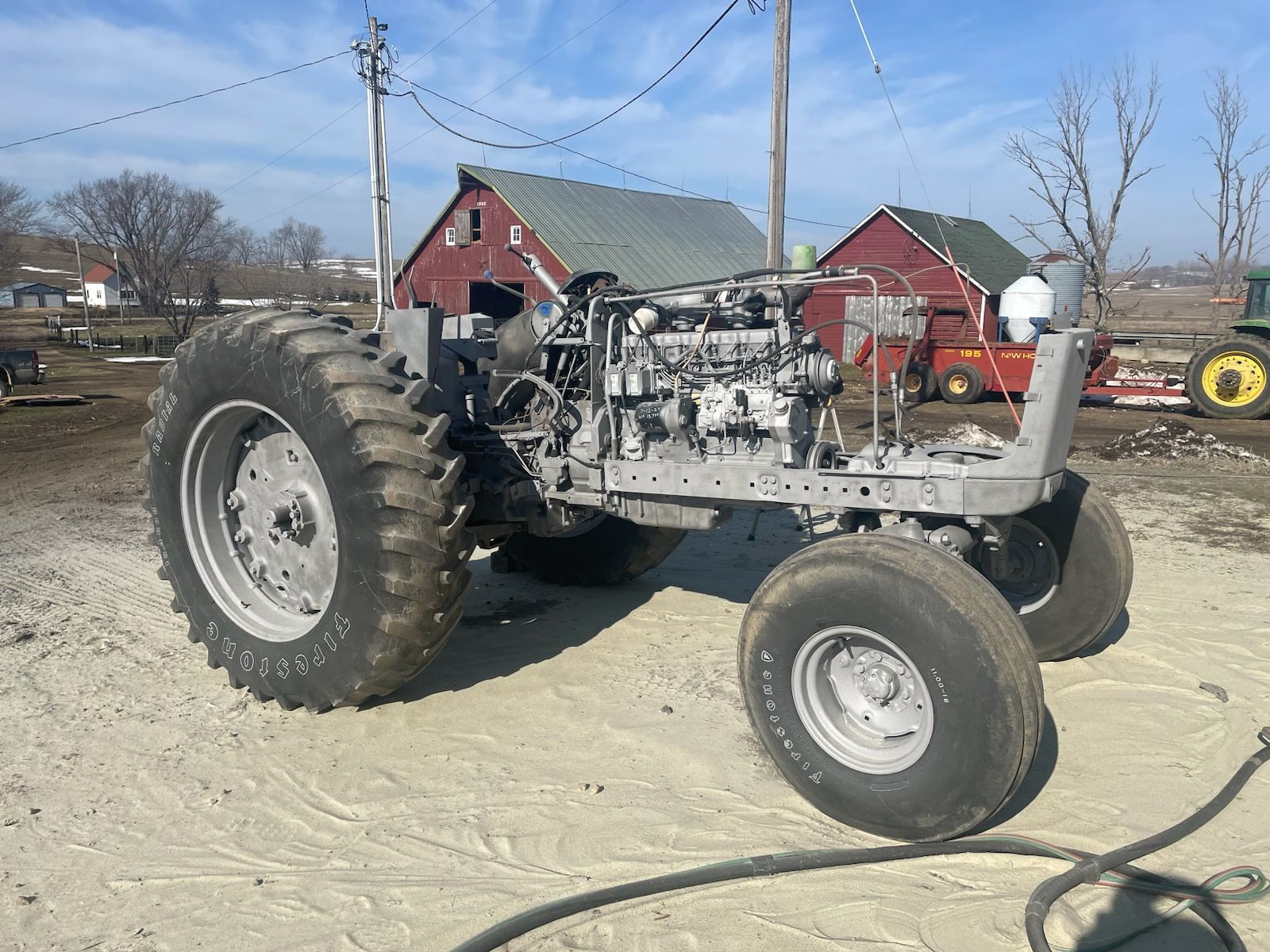 A disassembled gray tractor chassis with large tires sitting on a dirt farm lot in front of red barns.