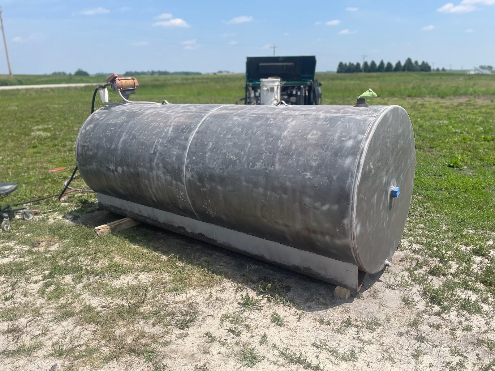 A dark cylindrical fuel tank sits on a metal stand in a grassy field with a pump unit behind it.