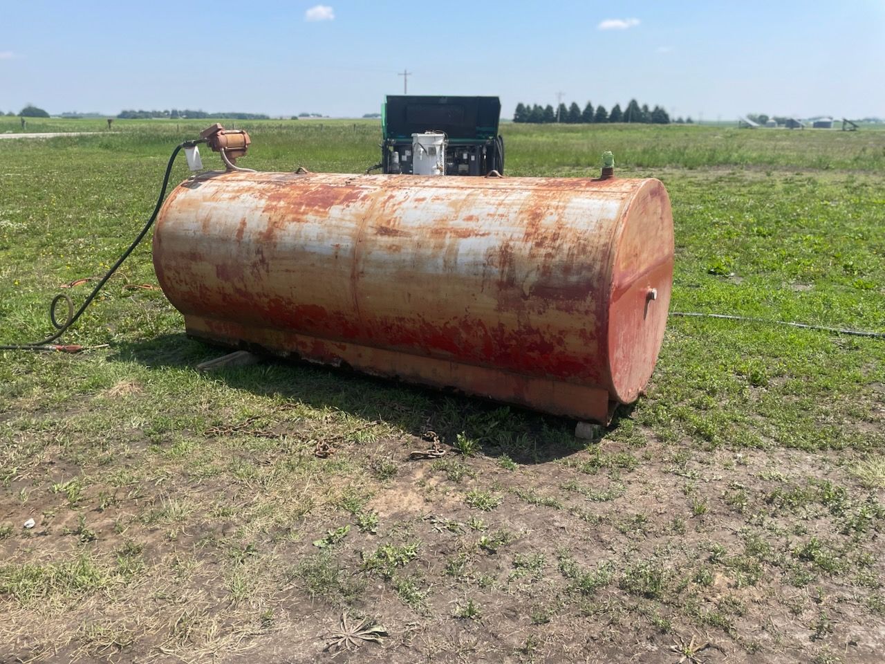 A rusty, horizontal metal fuel tank sitting in a grassy field under a clear blue sky.