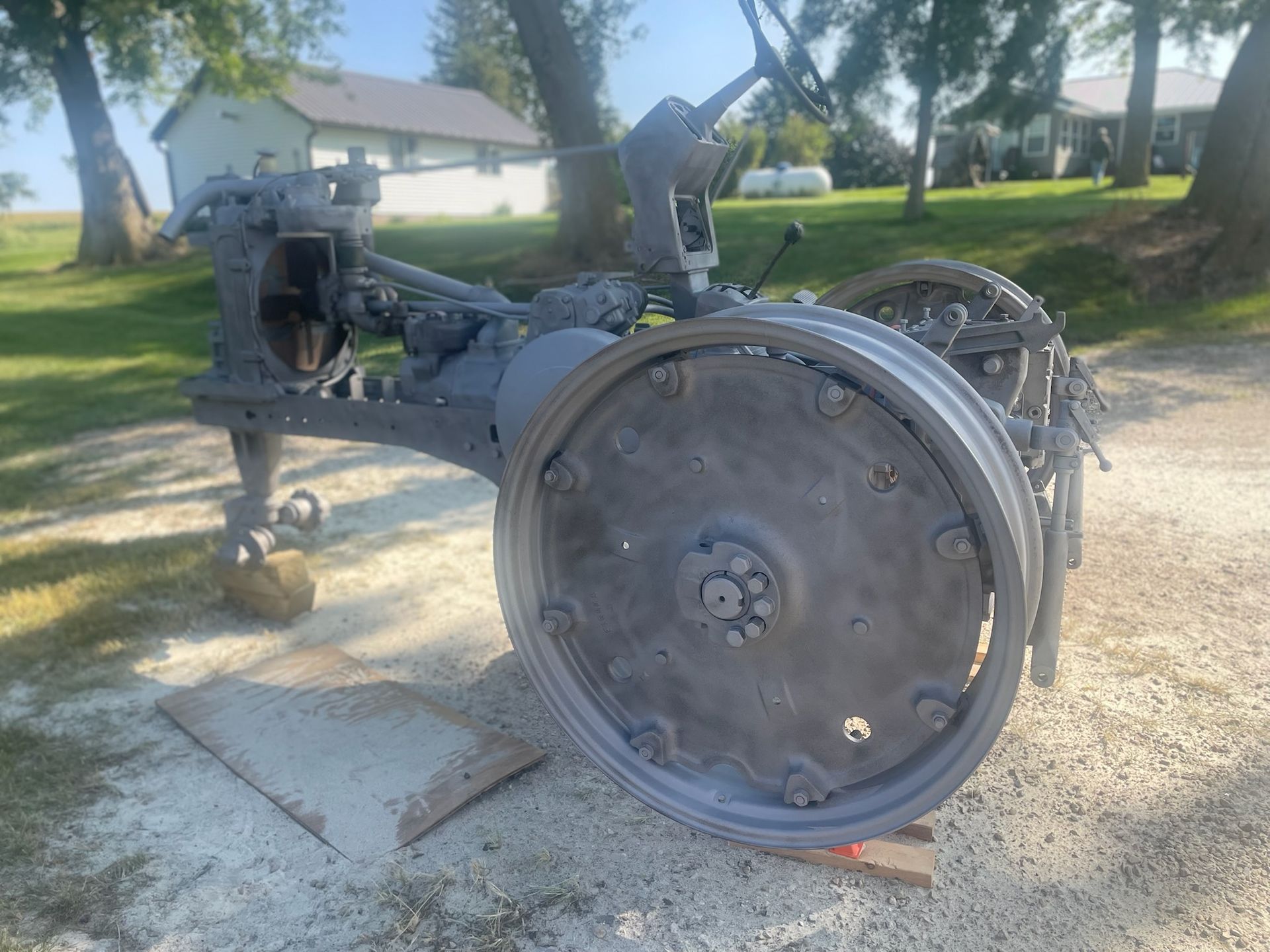 A vintage, gray-painted tractor frame with large steel wheels sits on a gravel drive near a house and trees.