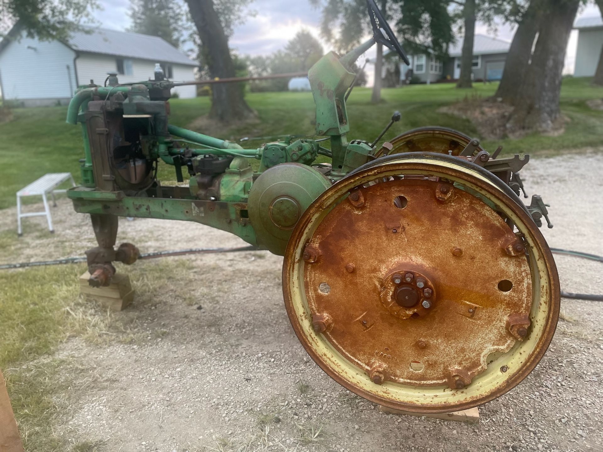 An antique green John Deere tractor sits on a gravel driveway near trees and a house, with a large, rusted rear wheel.