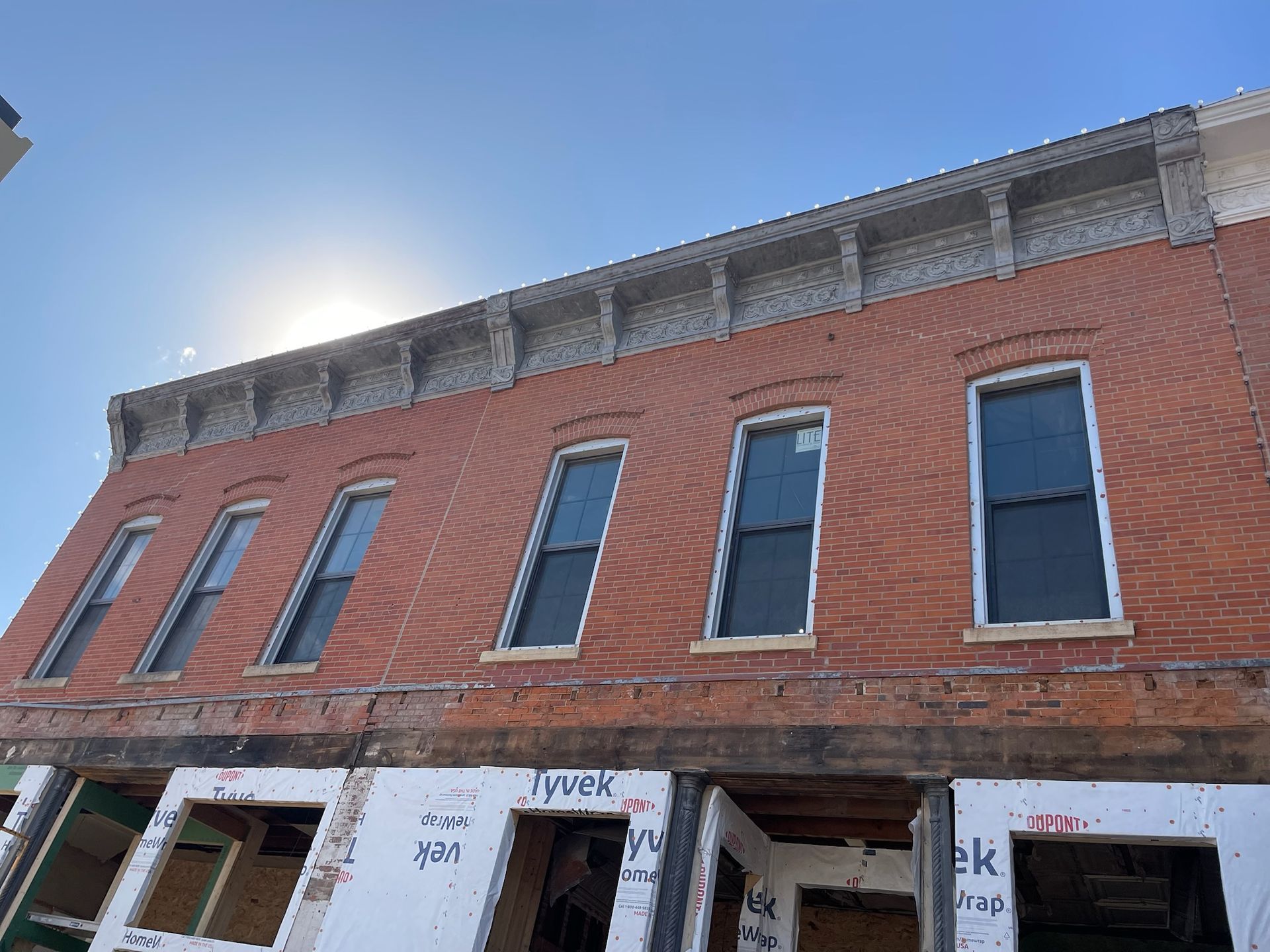 A brick two-story building under construction with a decorative cornice, dark windows, and Tyvek-covered storefronts.
