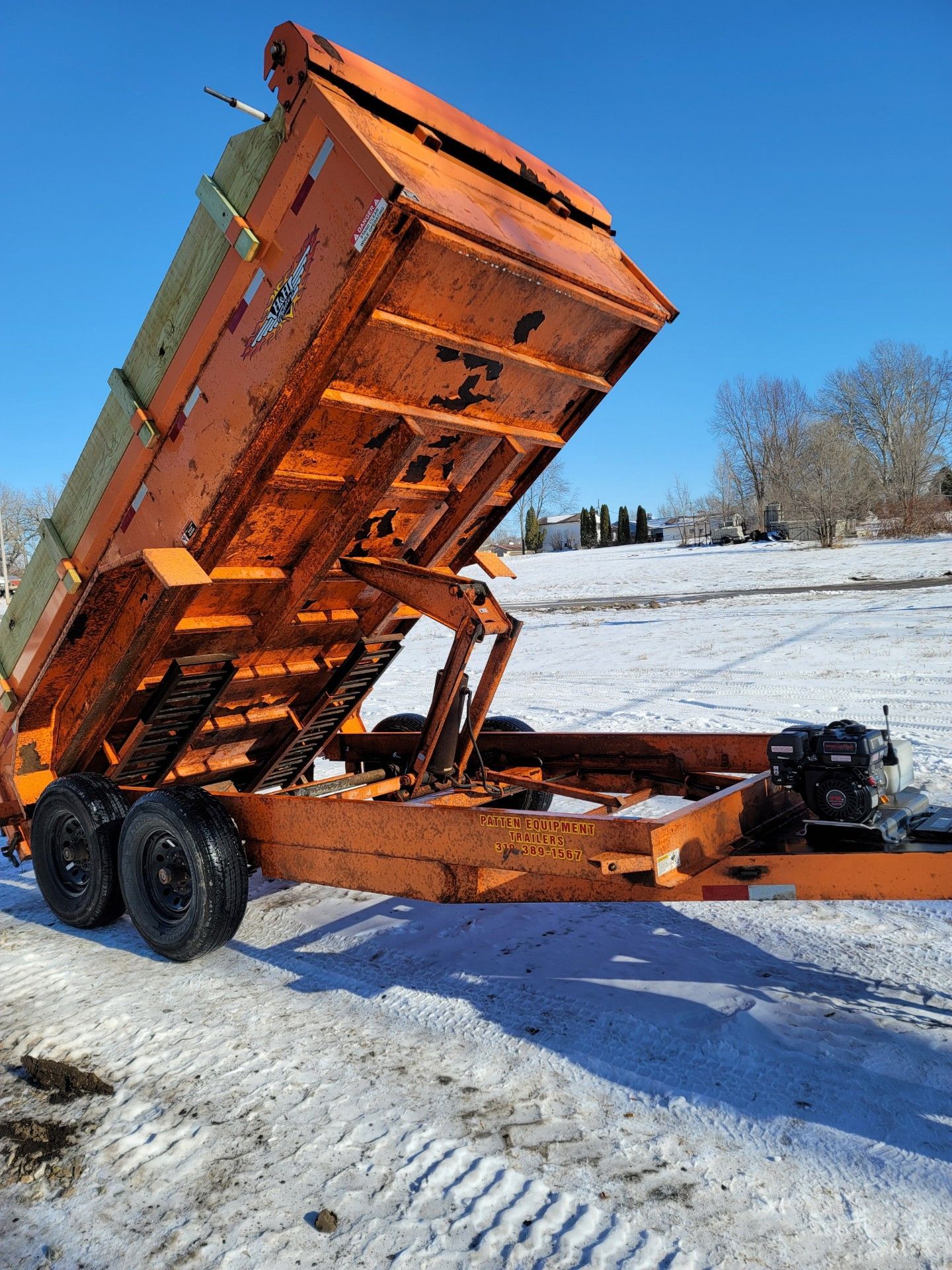 An orange dump trailer raised in the dumping position, parked on snow-covered ground under a clear blue sky.