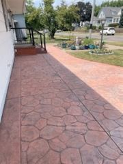 A stamped concrete walkway with a dark reddish-brown stone pattern leads from a house porch toward a suburban lawn.
