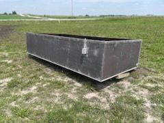 A large, rectangular dark metal livestock water tank sits on grass in an open field under a blue sky.