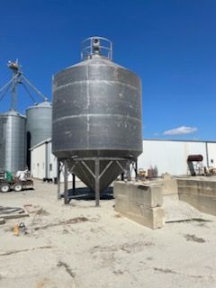 A metal feed or grain silo with a conical base stands in an outdoor industrial lot under a clear blue sky.