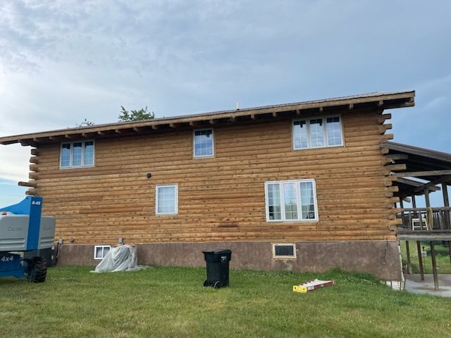A brown, two-story log cabin exterior featuring multiple white-framed windows and a small area of newer, lighter wood.