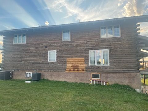Two-story log cabin exterior with multiple windows and a concrete foundation on a grassy lawn under a clear blue sky.