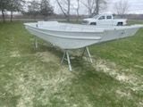 A white flat-bottom boat sits on metal stands in a grassy field with a white pickup truck parked in the background.
