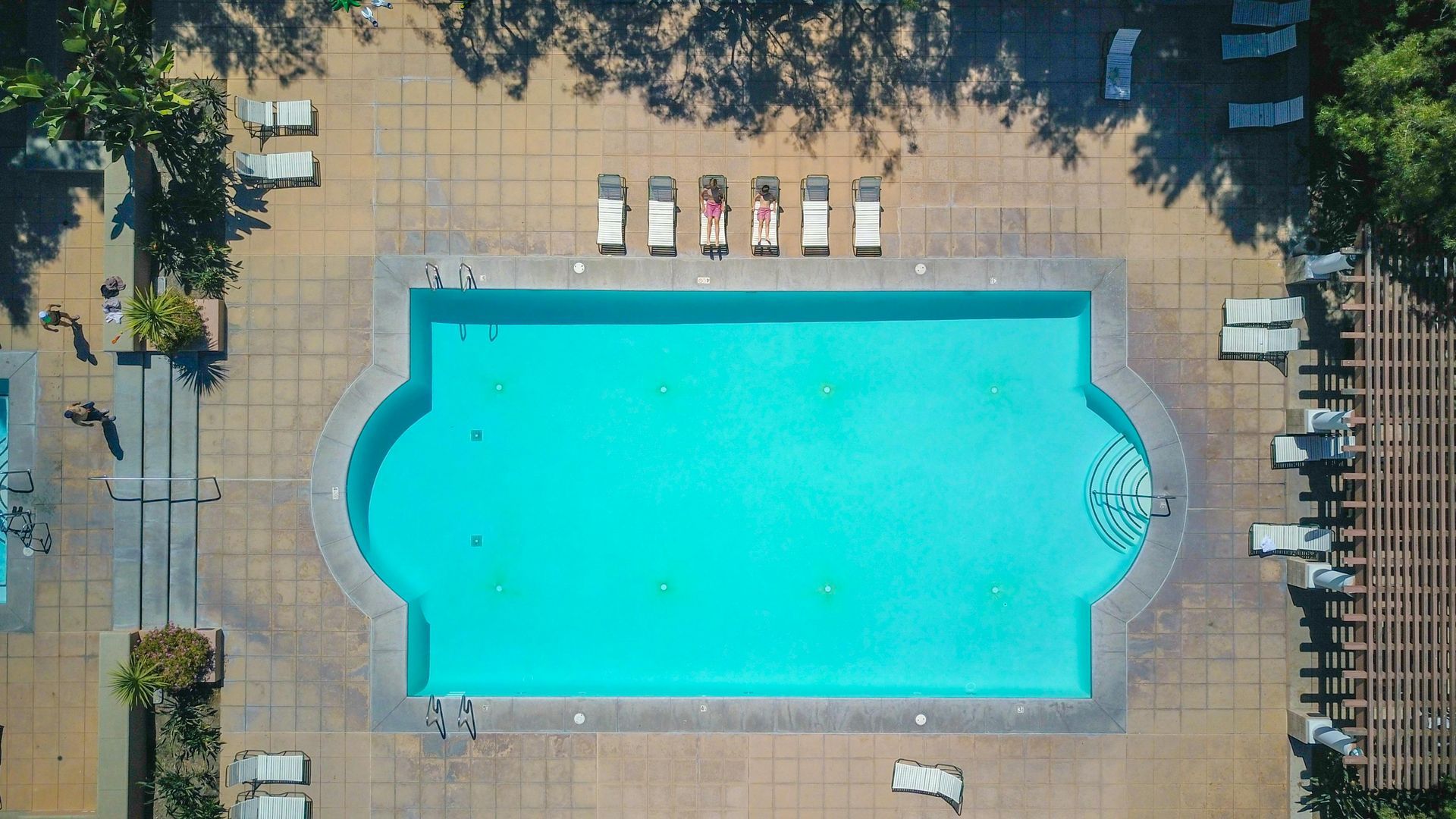 A top-down view of a rectangular, turquoise swimming pool with rounded corners, surrounded by lounge chairs and patio.