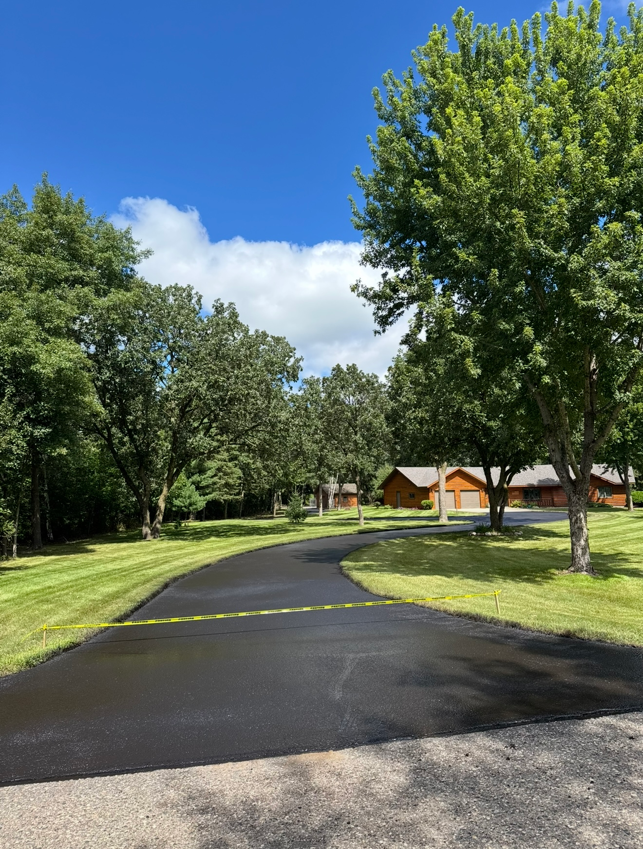 Curved wet asphalt path through a green park with trees and houses under a blue sky