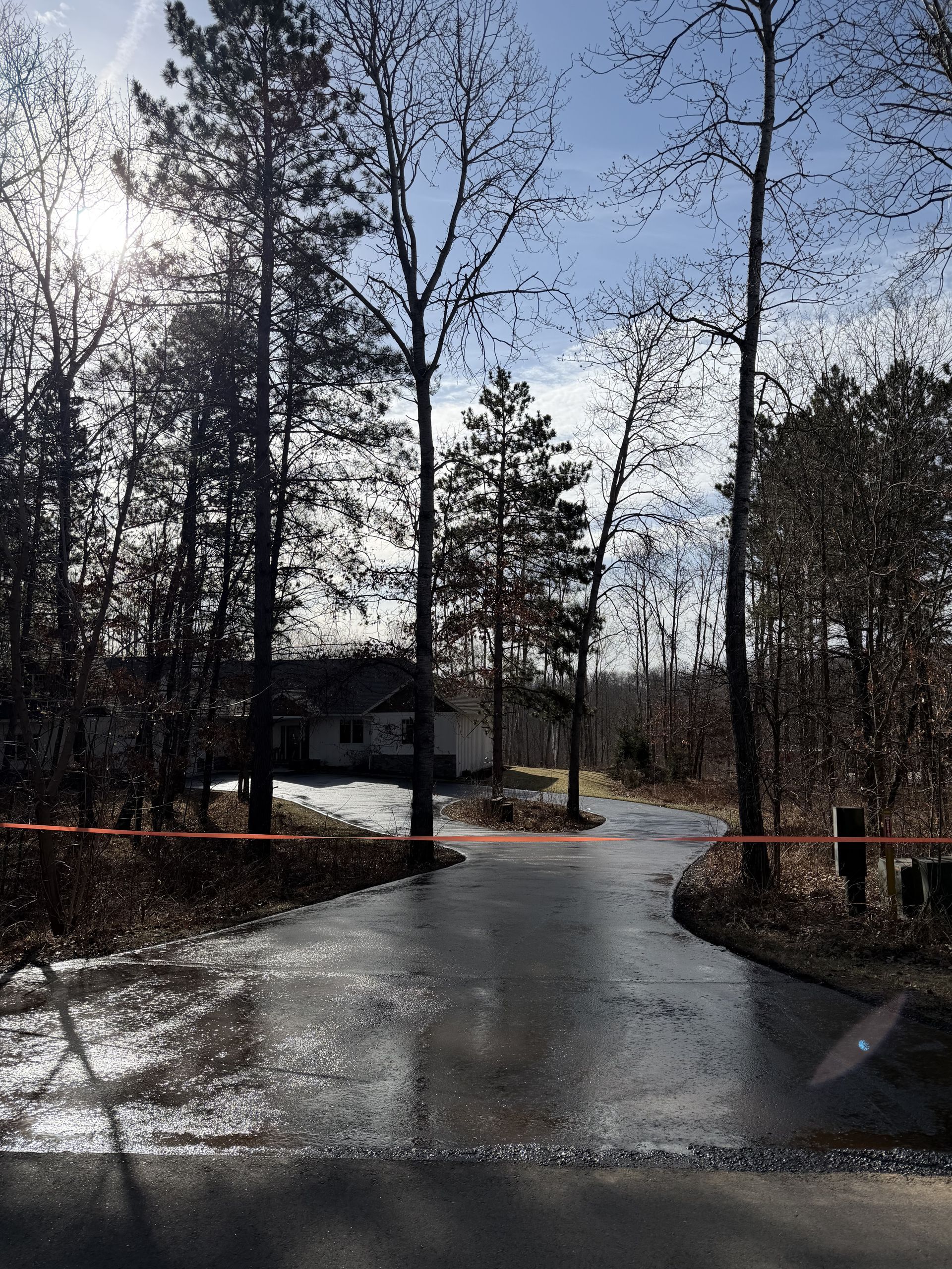 Wet driveway in a wooded area with a house in the background and cloudy sky.