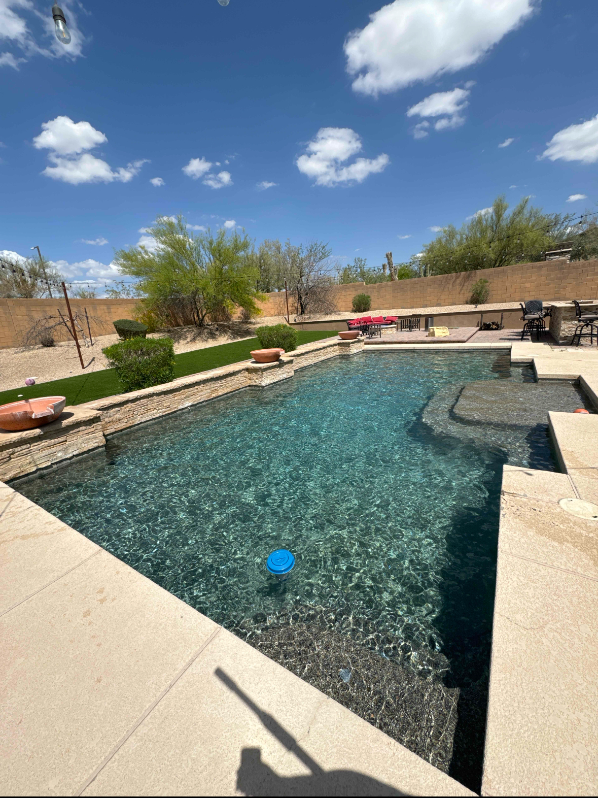 Backyard pool on a sunny day, clear blue water, beige stone surround, blue sky with clouds.