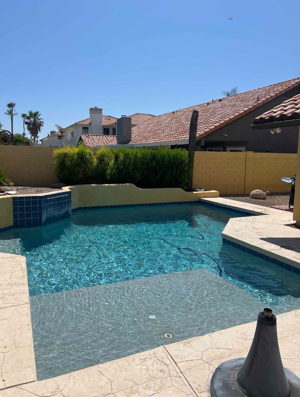 Swimming pool with blue water surrounded by beige concrete and a yellow wall, under a clear sky.