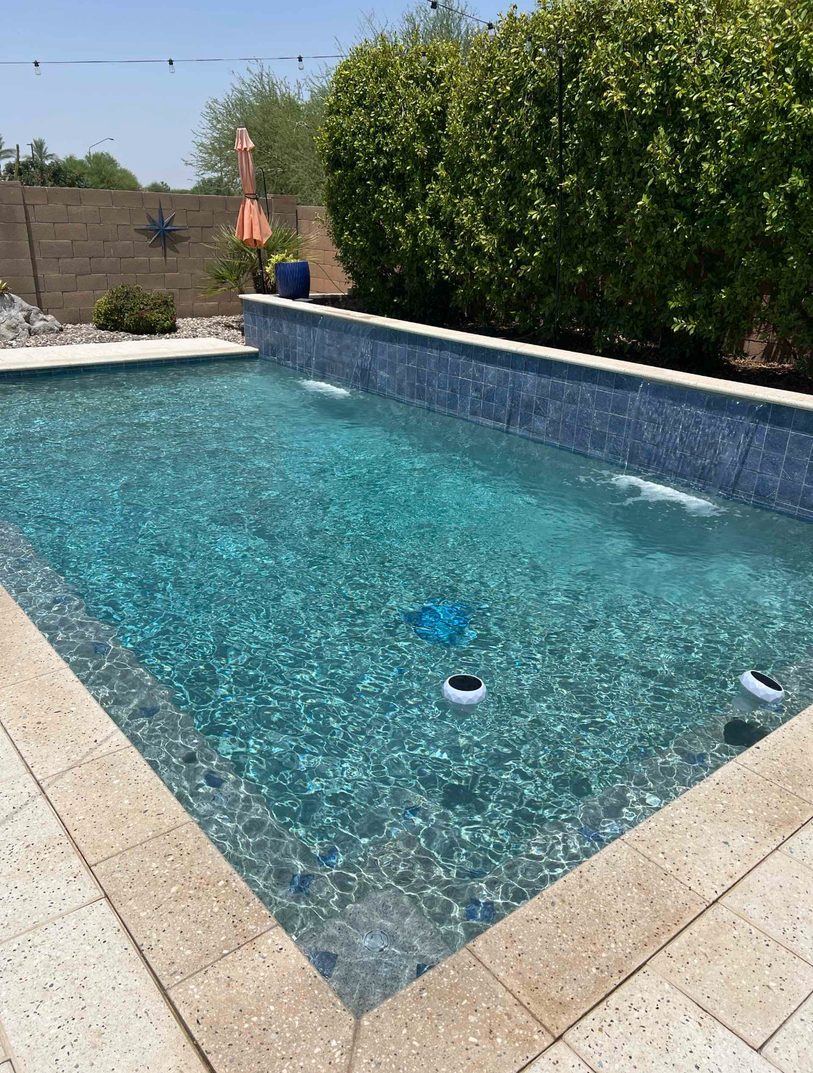 Rectangular turquoise pool with blue tile trim, surrounded by light stone, next to a green bush.
