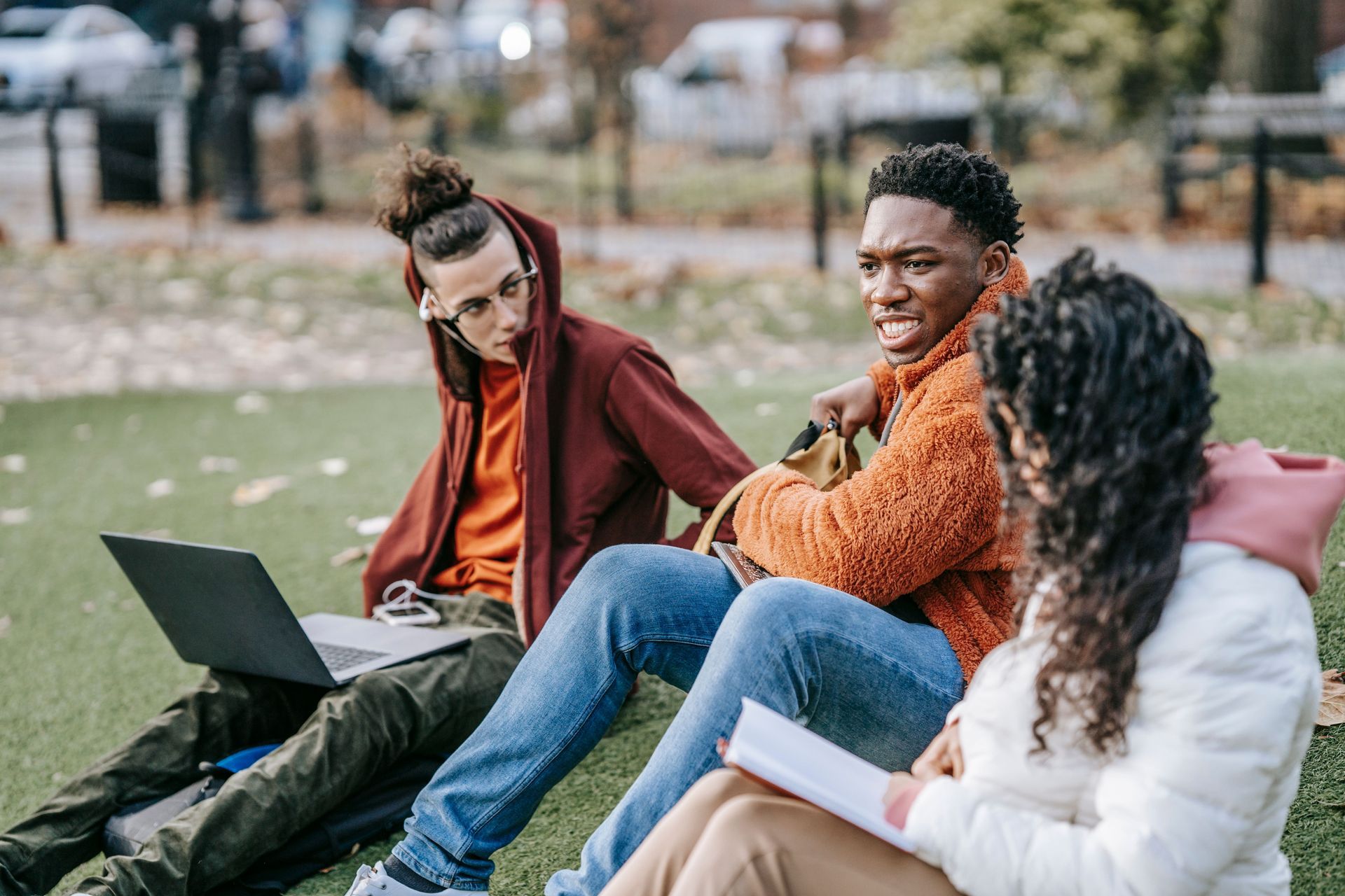 Three people sitting on grass outdoors, talking with a laptop open between them