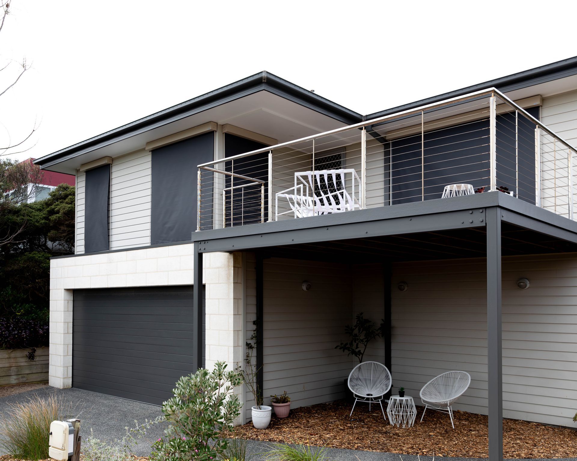 Two-story modern home with gray and white siding, balcony, and garage. Two chairs sit on the ground level.