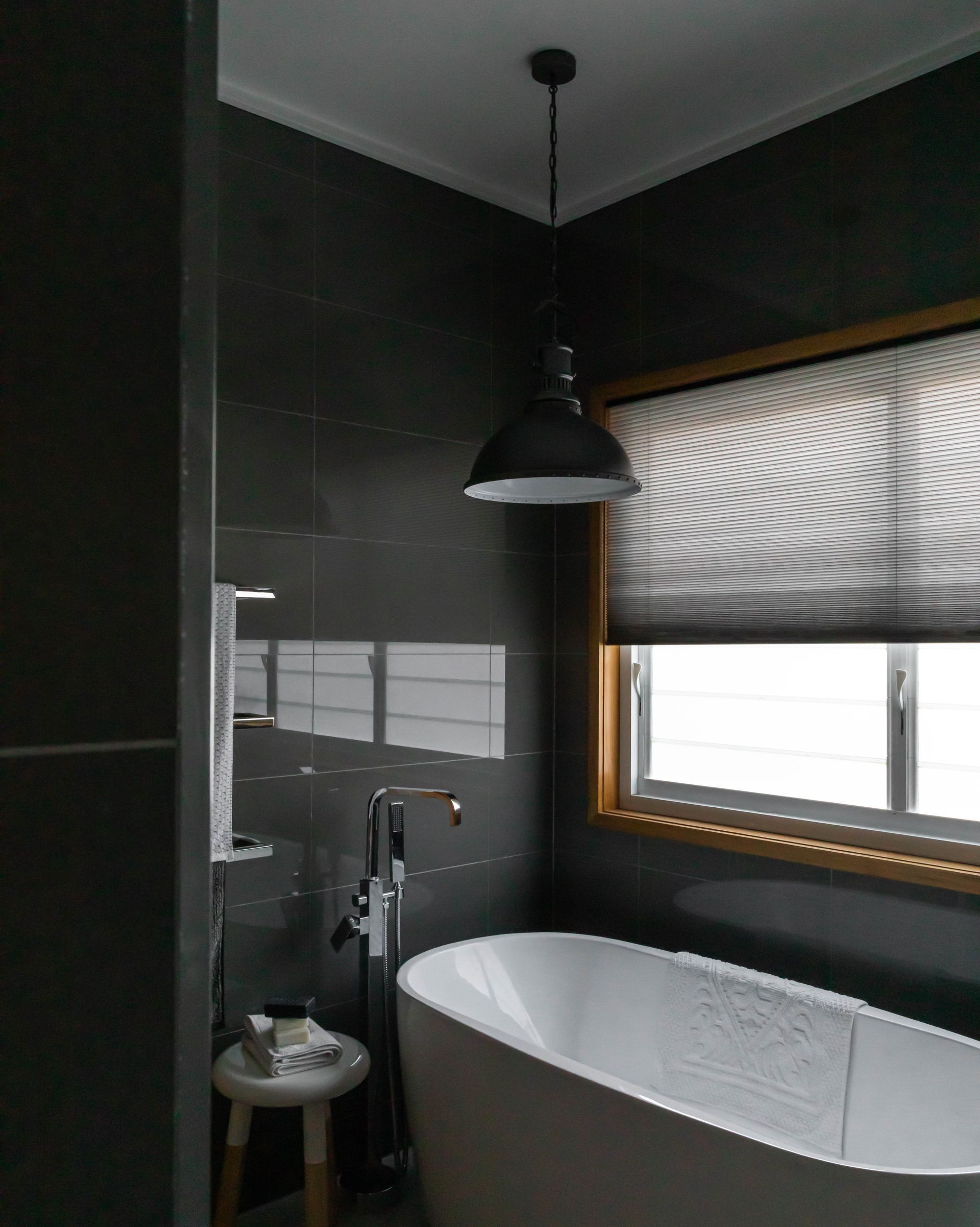 Bathroom with dark gray tile walls, white tub, window with shade, and pendant light.