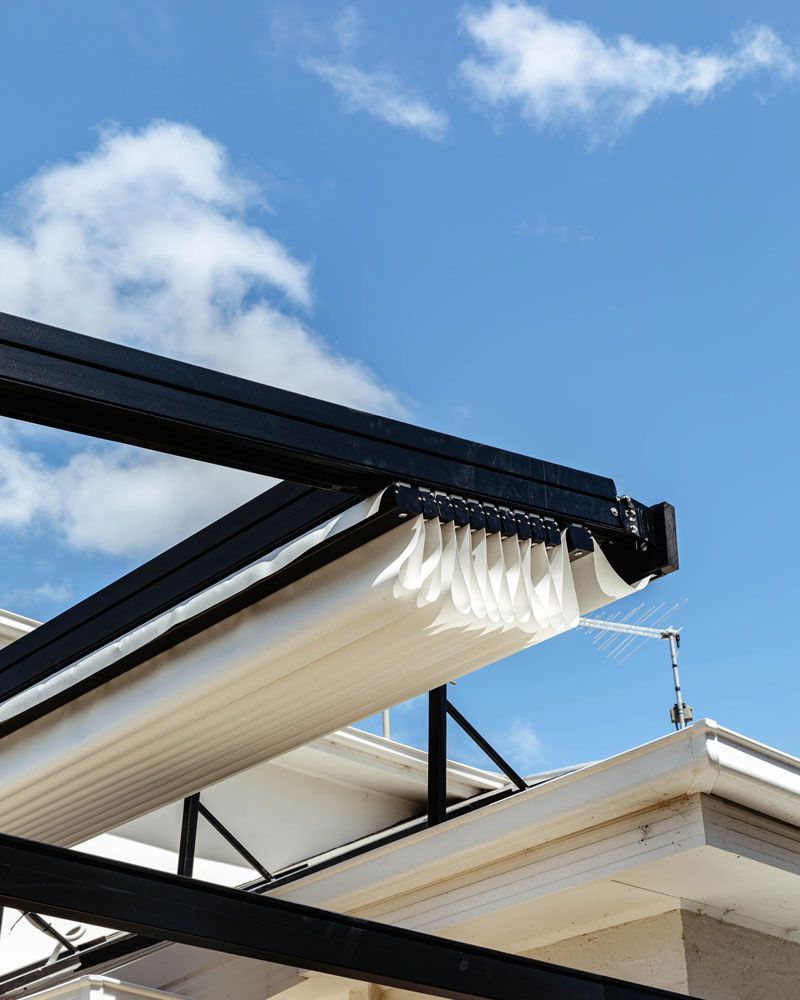 Black pergola with cream-colored retractable awning against a bright blue sky with scattered clouds.