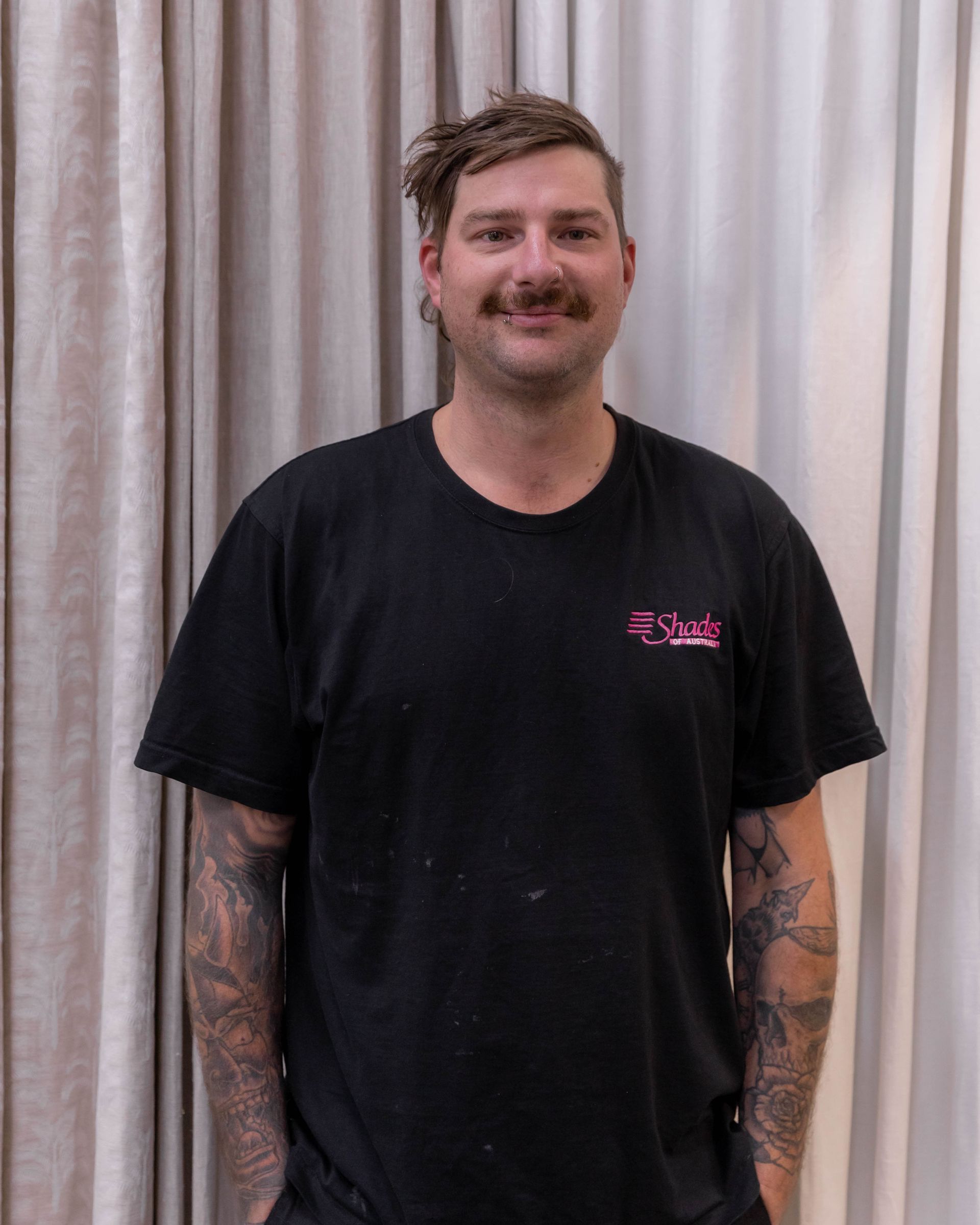 Man with tattoos wearing black t-shirt, arms in pockets, standing in front of neutral curtains.