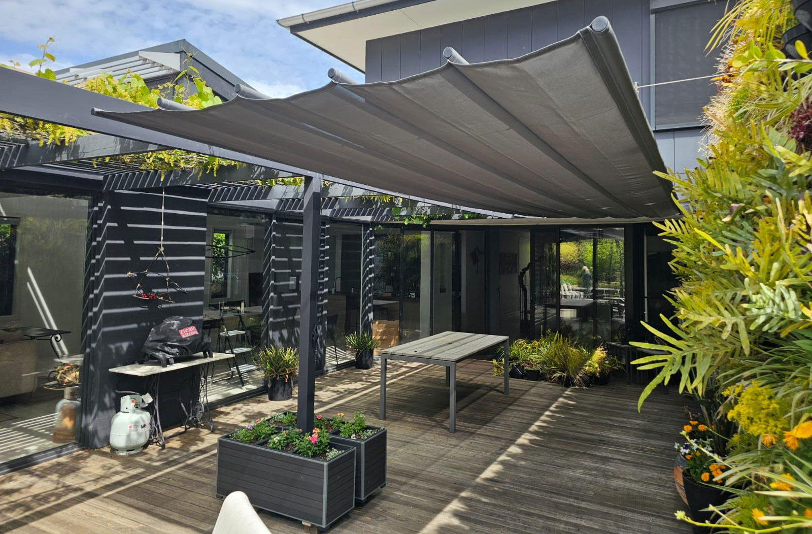 Patio with retractable gray awning over a dining table. Plants, dark wood pergola, and grilling area visible.