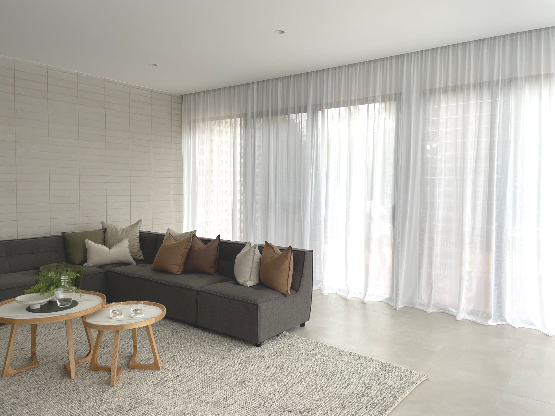 Living room with gray sectional sofa, white sheer curtains, wooden coffee tables, and a textured rug.