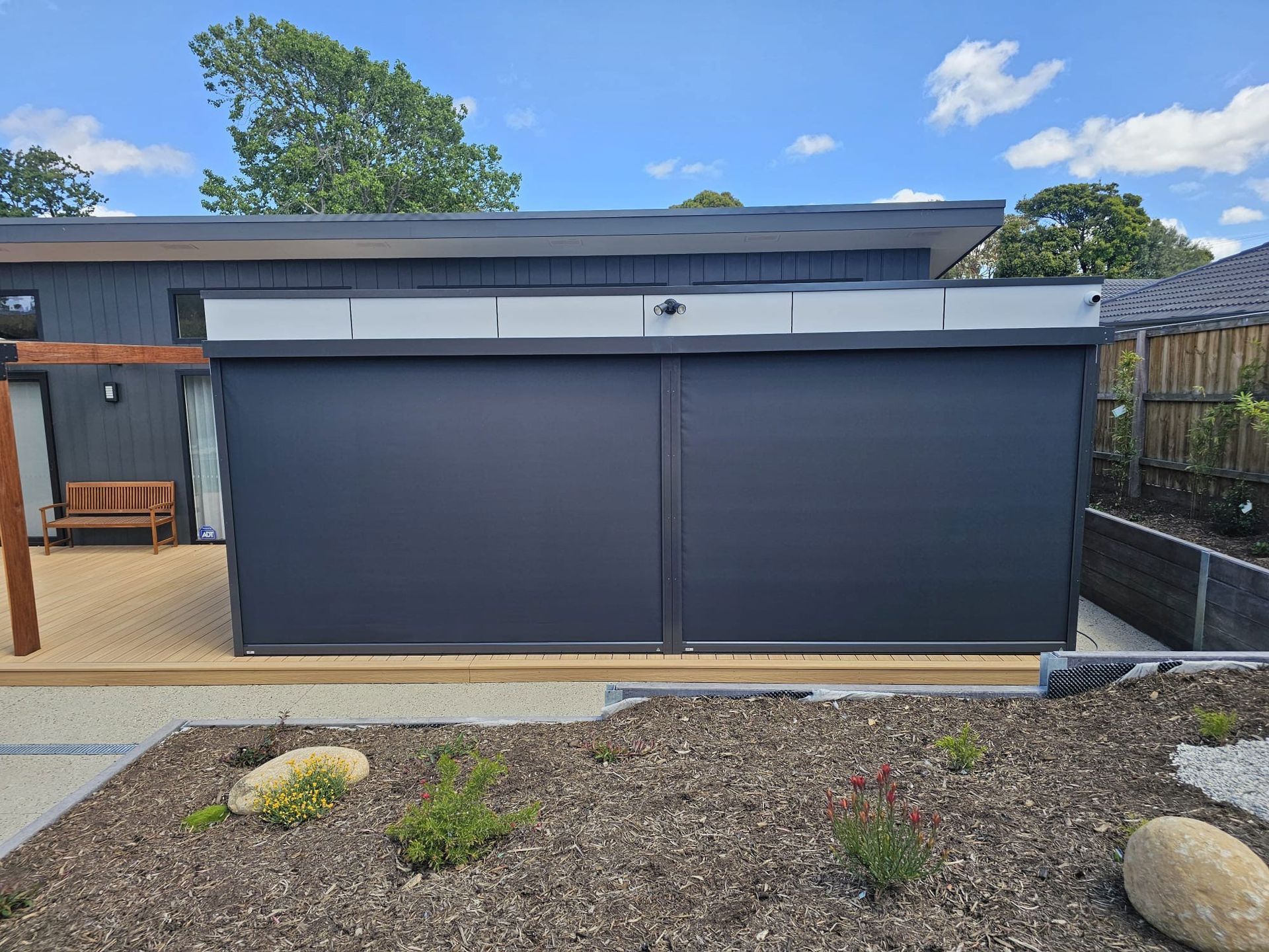 Black outdoor blinds on a modern building, with a wooden deck and garden in front.