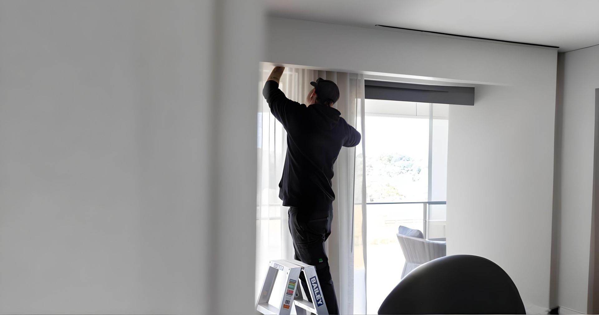 Man on a stepladder installing curtains near a window. He's wearing a dark jacket, working in a room.