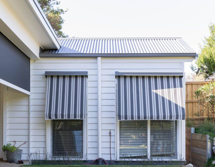 Two windows with blue and white striped awnings on a white building with a corrugated metal roof.